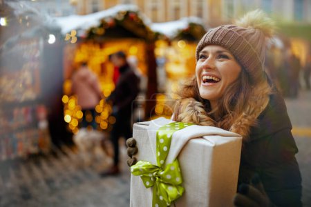 Photo for Happy modern 40 years old woman in green coat and brown hat at the winter fair in the city with christmas gift. - Royalty Free Image
