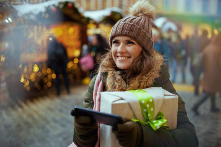 Téléchargez les photos : Heureuse femme moderne en manteau vert et chapeau brun à la foire de Noël dans la ville en utilisant smartphone. - en image libre de droit