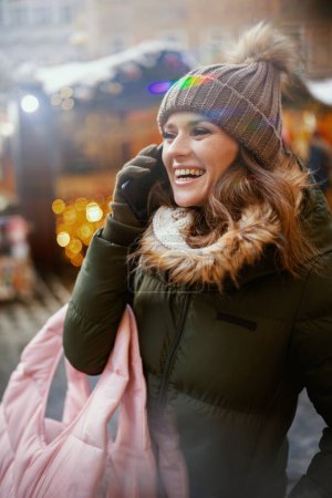Téléchargez les photos : Femme moderne souriante en manteau vert et chapeau marron à la foire d'hiver en ville parlant sur un smartphone. - en image libre de droit