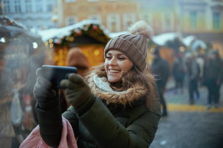 Photo for Happy modern middle aged woman in green coat and brown hat at the christmas fair in the city using video chat. - Royalty Free Image