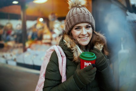 Photo for Smiling modern woman in green coat and brown hat at the winter fair in the city with Christmas beverage. - Royalty Free Image
