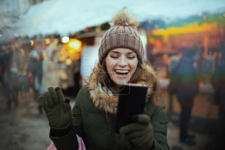 Foto de Sonriente mujer moderna de mediana edad en abrigo verde y sombrero marrón en la feria de invierno en la ciudad teniendo videollamada. - Imagen libre de derechos