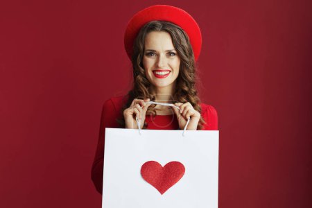 Téléchargez les photos : Joyeux Valentin. souriant élégante femme d'âge moyen en robe rouge et béret avec sac à provisions avec coeur isolé sur rouge. - en image libre de droit
