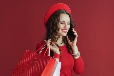 Foto de Feliz San Valentín. mujer moderna feliz en vestido rojo y boina sobre fondo rojo con bolsas de compras hablando en un teléfono inteligente. - Imagen libre de derechos
