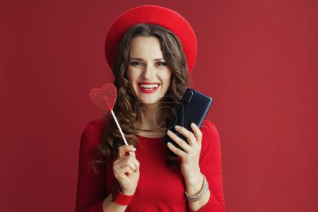 Foto de Feliz San Valentín. sonriente mujer moderna de 40 años en vestido rojo y boina con teléfono inteligente y caramelo en forma de corazón en palo aislado en rojo. - Imagen libre de derechos