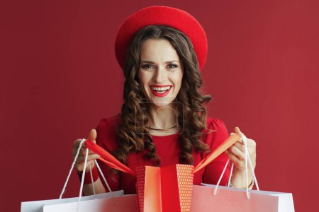 Téléchargez les photos : Joyeux Valentin. Portrait de femme moderne heureuse avec de longs cheveux ondulés en robe rouge et béret sur fond rouge avec des sacs à provisions. - en image libre de droit