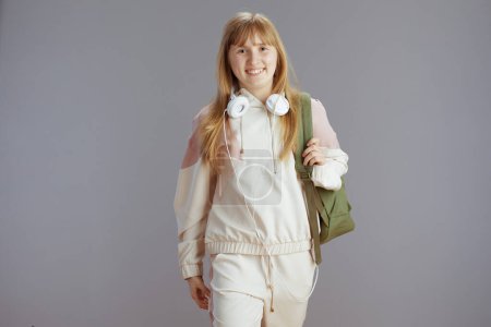 Téléchargez les photos : Portrait d'adolescente tendance souriante en survêtement beige avec sac à dos et écouteurs contre gris. - en image libre de droit