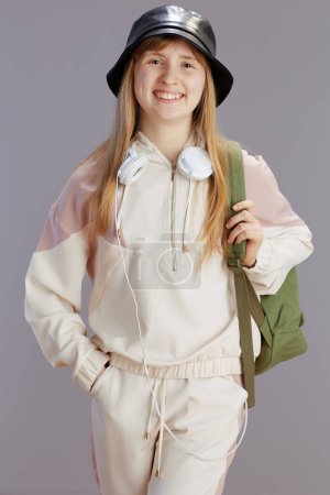 Photo for Portrait of happy modern school girl in beige tracksuit with backpack, headphones and hat isolated on grey. - Royalty Free Image