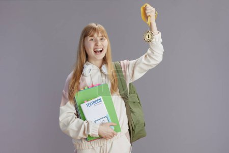 Téléchargez les photos : Jeune femme moderne souriante en survêtement beige avec sac à dos, classeurs, écouteurs, médailles et manuels isolés sur gris. - en image libre de droit
