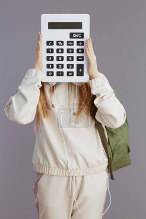 Photo for Modern teenage girl in beige tracksuit with backpack and calculator isolated on grey background. - Royalty Free Image