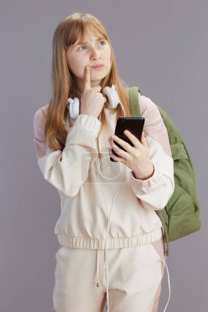 Photo for Pensive trendy school girl in beige tracksuit with backpack, headphones and smartphone isolated on grey. - Royalty Free Image