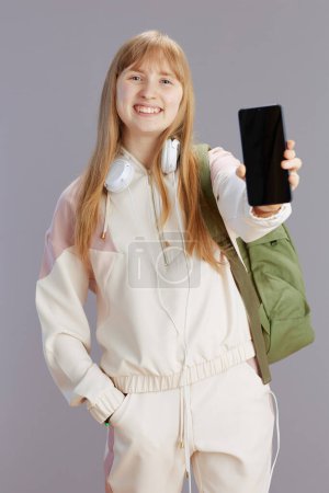 Photo for Portrait of happy modern school girl in beige tracksuit with backpack and headphones showing smartphone blank screen isolated on grey. - Royalty Free Image