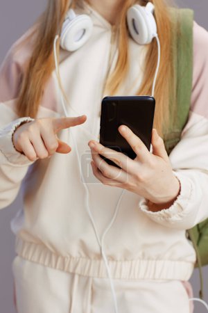 Photo for Closeup on teen girl in beige tracksuit with backpack and headphones using smartphone app against grey. - Royalty Free Image