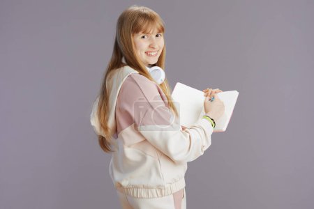 Foto de Retrato de chica de moda feliz en chándal beige con libros de trabajo y auriculares escribiendo contra gris. - Imagen libre de derechos