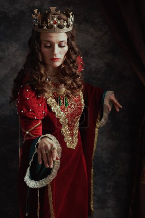 Photo for Medieval queen in red dress with crown making reverence on dark gray background. - Royalty Free Image