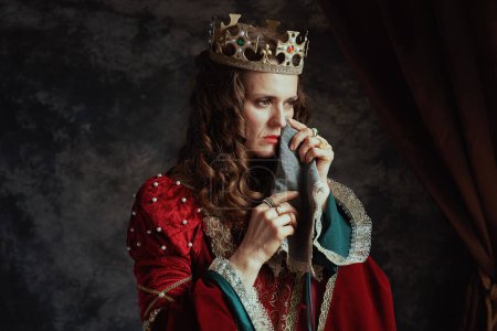Téléchargez les photos : Triste reine médiévale en robe rouge avec mouchoir et couronne pleurant sur fond gris foncé. - en image libre de droit