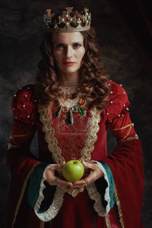 Téléchargez les photos : Reine médiévale en robe rouge avec pomme verte et couronne sur fond gris foncé. - en image libre de droit