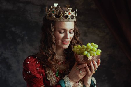 Téléchargez les photos : Reine médiévale souriante en robe rouge avec assiette de raisins et couronne sur fond gris foncé. - en image libre de droit