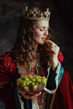Foto de Reina medieval sonriente en vestido rojo con plato de uvas y corona sobre fondo gris oscuro. - Imagen libre de derechos
