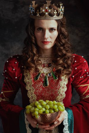 Foto de Reina medieval en vestido rojo con plato de uvas y corona sobre fondo gris oscuro. - Imagen libre de derechos