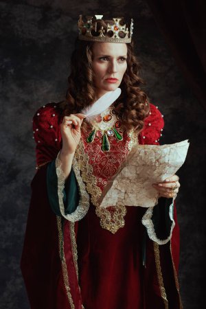 Foto de Pensativa reina medieval en vestido rojo con pergamino y corona sobre fondo gris oscuro. - Imagen libre de derechos