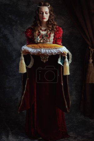 Foto de Retrato completo de la reina medieval en vestido rojo con corona sobre almohada sobre fondo gris oscuro. - Imagen libre de derechos