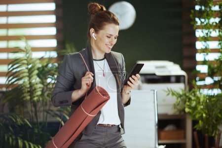 Photo for Sustainable workplace. smiling modern business woman at work with wireless headphones and yoga mat using smartphone applications. - Royalty Free Image