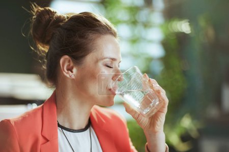 Foto de Lugar de trabajo sostenible. mujer de negocios moderna en moderna oficina verde en una chaqueta roja con taza de agua. - Imagen libre de derechos