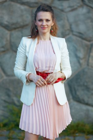 Foto de Mujer elegante en vestido rosa y chaqueta blanca en la ciudad contra la pared de piedra. - Imagen libre de derechos