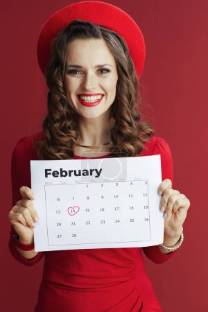 Téléchargez les photos : Joyeux Valentin. heureux élégant 40 ans femme en robe rouge et béret isolé sur fond rouge avec calendrier Février. - en image libre de droit