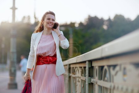 Foto de Sonriente mujer moderna en vestido rosa y chaqueta blanca en la ciudad hablando en un teléfono inteligente en el puente. - Imagen libre de derechos