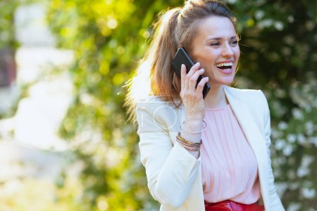 Téléchargez les photos : Femme souriante à la mode en robe rose et veste blanche dans la ville parlant sur un smartphone. - en image libre de droit