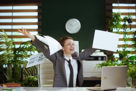 Photo for New job. smiling modern woman worker in modern green office in grey business suit with laptop throwing documents. - Royalty Free Image