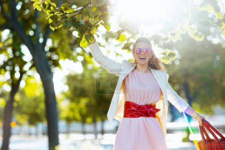 Téléchargez les photos : Femme d'âge moyen moderne souriante en robe rose et veste blanche dans la ville avec des lunettes de soleil. - en image libre de droit