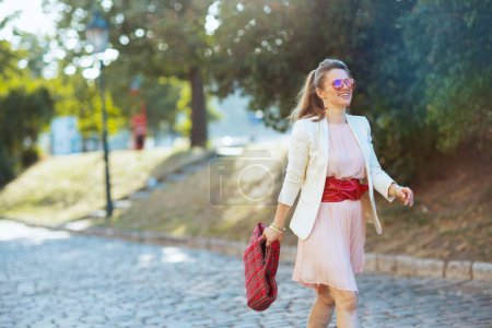 Téléchargez les photos : Souriant élégant femme d'âge moyen en robe rose et veste blanche dans la ville avec sac rouge et lunettes de soleil marche. - en image libre de droit