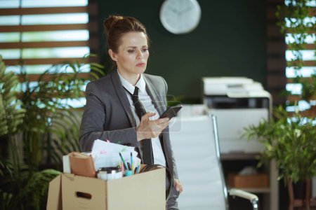 Téléchargez les photos : Un nouveau boulot. coûteuse femme d'âge moyen moderne employée dans un bureau vert moderne en costume d'affaires gris avec des effets personnels dans une boîte en carton et un smartphone. - en image libre de droit