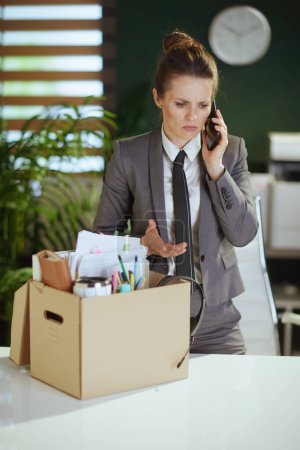 Photo for New job. stressed modern woman worker in modern green office in grey business suit with personal belongings in cardboard box speaking on a smartphone. - Royalty Free Image