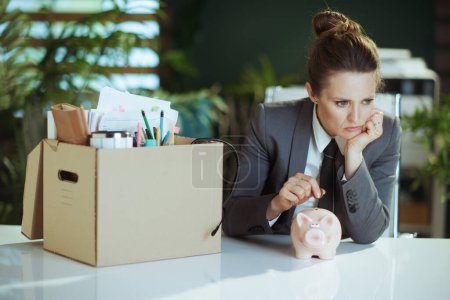 Photo for New job. pensive modern 40 years old woman employee in modern green office in grey business suit with personal belongings in cardboard box putting coin into piggy bank. - Royalty Free Image