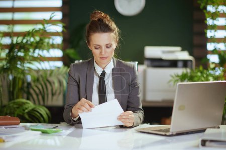 Téléchargez les photos : Un nouveau boulot. coûteuse femme moderne de 40 ans employée dans un bureau vert moderne en costume d'affaires gris avec lettre d'ouverture pour ordinateur portable. - en image libre de droit