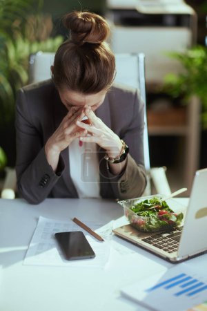 Photo for Sustainable workplace. stressed modern middle aged business woman in a grey business suit in modern green office with laptop eating salad and using smartphone app. - Royalty Free Image