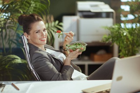 Photo for Sustainable workplace. happy modern 40 years old woman worker in a grey business suit in modern green office with laptop eating salad. - Royalty Free Image
