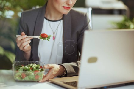 Photo for Sustainable workplace. Closeup on middle aged woman employee in a grey business suit in green office with laptop eating salad. - Royalty Free Image