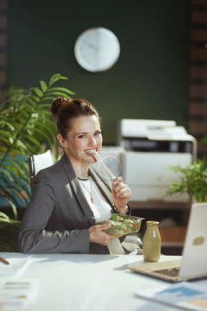 Foto de Lugar de trabajo sostenible. feliz mujer de mediana edad moderna empleada en un traje de negocios gris en la oficina verde moderna con portátil comer ensalada. - Imagen libre de derechos