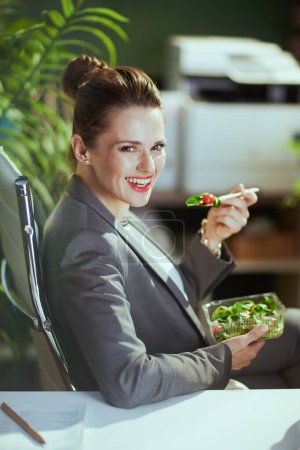 Foto de Lugar de trabajo sostenible. feliz mujer de mediana edad moderna empleado en un traje de negocios gris en la oficina verde moderna comer ensalada. - Imagen libre de derechos
