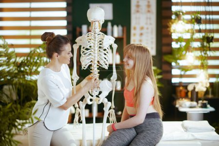 Photo for Healthcare time. happy female medical massage therapist in massage cabinet with teenage client and skeleton. - Royalty Free Image