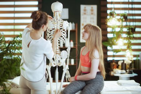 Photo for Healthcare time. concerned massage therapist woman in massage cabinet with teenage client and skeleton. - Royalty Free Image