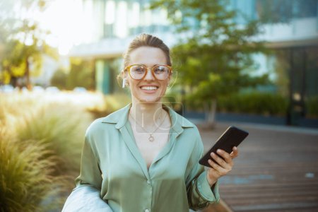 Photo for Portrait of happy modern woman worker near office building in green blouse and eyeglasses with smartphone. - Royalty Free Image