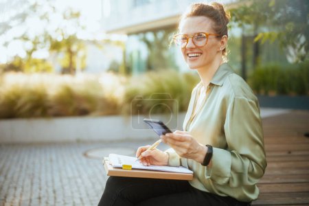 Photo for Smiling modern female worker in business district in green blouse and eyeglasses with smartphone, documents and folder. - Royalty Free Image