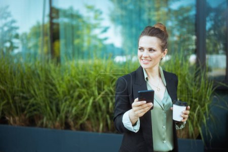 Téléchargez les photos : Travailleuse moderne souriante près du centre d'affaires en veste noire avec tasse de café à l'aide d'un smartphone. - en image libre de droit