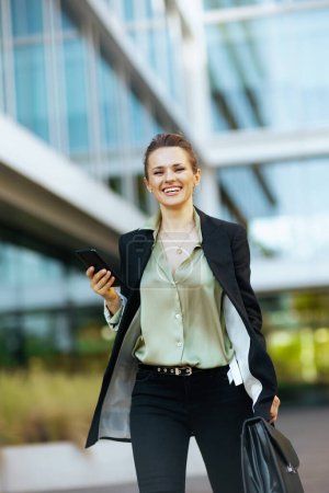 Photo for Happy modern middle aged woman worker in business district in black jacket with briefcase using smartphone and walking. - Royalty Free Image